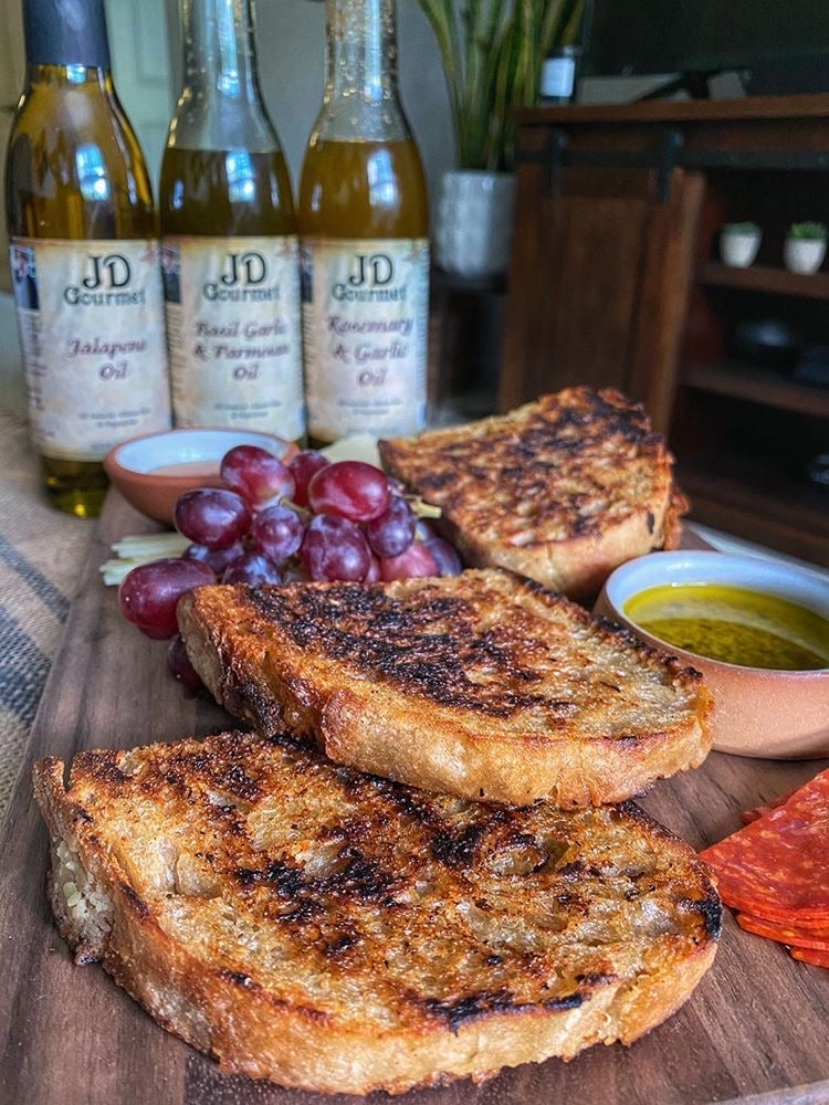 Grilled sourdough slices beside a bowl of herbed olive oil, with JD Gourmet Basil Garlic & Parmesan Oil on the board behind.